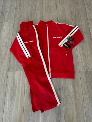 Rött tracksuit från Palm Angels - Snyggt rött tracksuit från Palm Angels med vita ränder längs ärmar och ben. Hela settet inkluderas. Köp dina sommarkläder här! Jag har flera färger. Tar du fler så får du en fin rabbat. Fraktas omedelbart vid köp. 