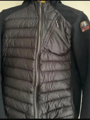 Svart pufferjacka från Parajumpers - Snygg svart pufferjacka från Parajumpers med quiltad framsida och rygg, huva och dragkedja framtill. Jackan har en bröstficka med dragkedja och patch med logga på ärmen. Materialmix med glansig yta och stickade partier på ärmar och sidor.