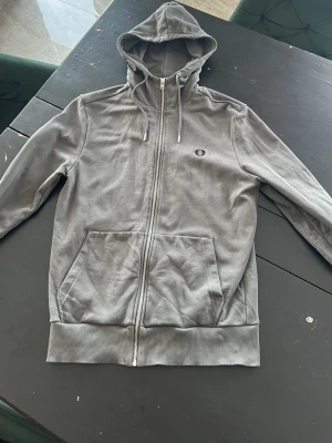 Grå zip-hoodie från Fred Perry - Snygg grå hoodie med dragkedja från Fred Perry. Klassisk design med huva och dragsko, två stora fickor framtill och broderad logga på bröstet. Perfekt för en avslappnad streetwear-look.