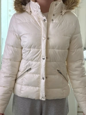 Vit pufferjacka med fuskpäls - Snygg vit pufferjacka med huva och fuskpälsdetalj. Jackan har dragkedja och tryckknappar framtill samt två snedställda fickor med dragkedja. Quiltad design som håller värmen och en klassisk stil. 