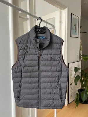 Grå väst från Polo Ralph Lauren - Snygg grå väst från Polo Ralph Lauren med diskret logga på bröstet. Västen har ståkrage, dragkedja framtill och två fickor med dragkedja. 