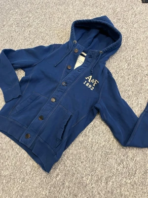 Blå hoodie från Abercrombie & Fitch - Snygg blå hoodie med knappar framtill och broderad A&F 1892-logga på bröstet. Tröjan har huva med dragsko, två fickor och ribbade muddar. Perfekt för en avslappnad och cool stil.