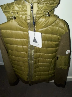 Moncler cardigan  - Säljer en olivgrön pufferjacka från Moncler med stickade ärmar och huva. Jackan har dragkedja framtill, justerbar huva med snörning och en ficka med lock på ena ärmen. Materialet är en mix av glansigt nylon och stickad ull på ärmarna. Moncler-logga på ärmen.