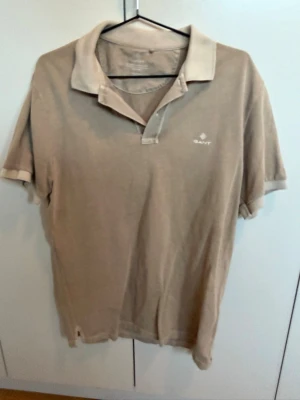 Beige kortärmad piké  från Gant - Säljer en beige piké  från Gant med klassisk krage och korta ärmar. T-shirten har en diskret logga på bröstet och är tillverkad i mjuk bomull. Perfekt för en avslappnad och stilren look.