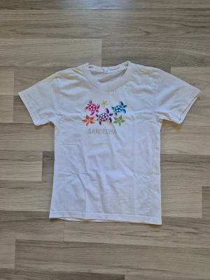 Vit t-shirt med färgglada sköldpaddor - Vit t-shirt med broderade sköldpaddor i olika färger och texten 'Sardegna' på bröstet. T-shirten har rund hals och korta ärmar, perfekt för dig som gillar unika och färgglada detaljer. Materialet känns som bomull och den har en normal passform.
