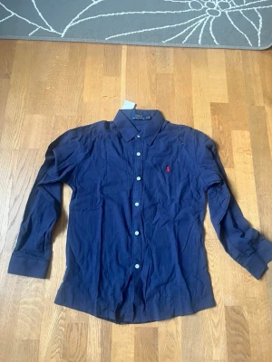 Mörkblå skjorta från Polo Ralph Lauren - Klassisk mörkblå skjorta från Polo Ralph Lauren med röd broderad logga på bröstet. Skjortan har långa ärmar, knappar framtill och är tillverkad i bomull. Perfekt för en stilren look.