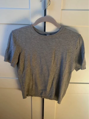Grå stickad topp - En enkel och stilren grå stickad topp från Divided med rund halsringning och korta ärmar. Toppen har ribbade detaljer vid hals, ärmslut och nederkant. Perfekt att matcha med jeans eller kjol för en clean look. Tröjan är lite i storleken så den är mer en xs.