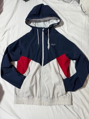 Hollister vindjacka i blått, vitt och rött - Snygg vindjacka från Hollister med färgblock i marinblått, vitt och rött. Jackan har huva med dragsko, hel dragkedja och två fickor framtill. Liten Hollister-logga på bröstet och elastisk nederkant. Perfekt för blåsiga dagar.