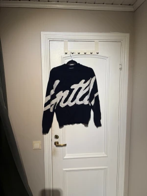 Svart stickad tröja med vit text från bershka - Cool svart stickad tröja med stor vit text i ett grafiskt mönster över hela framsidan. Tröjan har rund halsringning och långa ärmar. Perfekt för dig som gillar streetstyle och vill sticka ut lite extra.