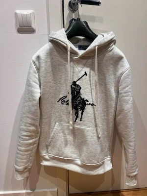 Polo Ralph Lauren hoodie - Snygg ljusgrå hoodie från Polo Ralph Lauren med stor svart polospelare-logga på bröstet. Klassisk känguruficka framtill, dragsko i huvan och mjukt material som känns skönt mot huden. Perfekt för en avslappnad och trendig stil. Storlek M men passar S