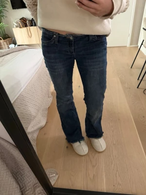 Blå bootcut jeans med råa kanter - Säljer ett par blå bootcut jeans med råa, fransiga benslut och låg midja. Jeansen har klassisk femficksdesign och är tillverkade i denim med lätt tvättad look. Perfekta för en avslappnad och trendig stil.