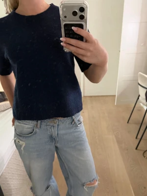 Mörkblå stickad t-shirttröja - Mörkblå stickad t-shirttröja med korta ärmar och rund halsringning. Tröjan har en enkel och stilren design med ribbstickade kanter vid hals, ärmslut och nederkant. Perfekt för lager-på-lager och passar till jeans eller kjol.