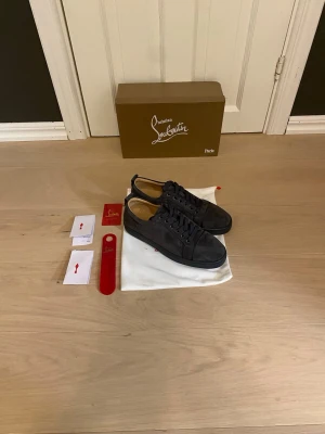 Mörkgrå sneakers från Christian Louboutin - Snygga mörkgrå sneakers från Christian Louboutin i mocka med klassisk låg siluett och matchande skosnören. Insidan är fodrad i ljust läder och skorna har diskreta detaljer som ger en exklusiv känsla. Perfekta för dig som gillar stilrena och lyxiga sneakers.
