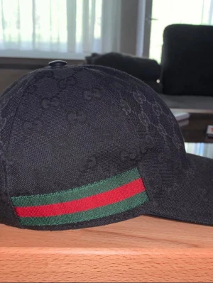 Svart keps från Gucci med GG-mönster - Snygg svart keps från Gucci med det klassiska GG-monogrammönstret över hela kepsen. Kepsen har en böjd skärm och justerbart band bak i läder. På sidan finns Guccis ikoniska gröna och röda rand. Perfekt accessoar för en trendig look.