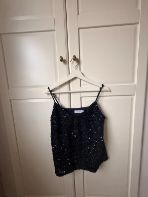Svart paljettlinne från Bik Bok - Glittrig svart linnetopp från Bik Bok med tunna axelband och paljettdetaljer. Perfekt för att sticka ut och ge outfiten extra bling. Snygg v-ringning och figurnära passform.