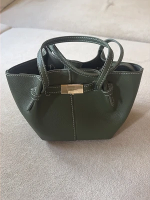 Olivgrön handväska med axelrem - Stilren olivgrön handväska i slätt konstläder med två handtag och avtagbar axelrem. Väskan har silverfärgat spänne framtill och vita kontrastsömmar som detalj. Perfekt storlek för det viktigaste och ger en trendig touch till din outfit.