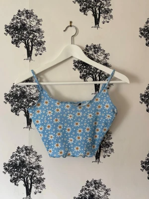 Blå blommig linnetopp med smala band - Söt blå linnetopp med smala axelband och blommigt mönster med vita och gula prästkragar. Toppen har vågig nederkant och är croppad, perfekt för sommarens varma dagar. Materialet är mjukt och stretchigt, vilket gör den bekväm att bära.