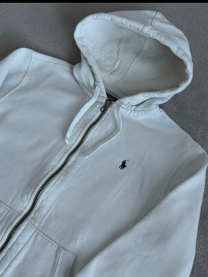 Vintage  Polo Ralph Lauren - Vit hoodie från Polo Ralph Lauren med dragkedja och klassisk logga broderad på bröstet. Modellen har huva med snören, ribbade muddar och två fickor framtill. Materialet är mjukt och bekvämt, perfekt för chill dagar.