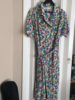 Blommig omlottklänning från Weinberg - Färgglad omlottklänning från Weinberg med blommigt mönster i blått, rosa, gult och grönt. Klänningen har korta ärmar, krage och knytskärp i midjan. Perfekt för dig som gillar retrovibbar och vill sticka ut med en unik look.