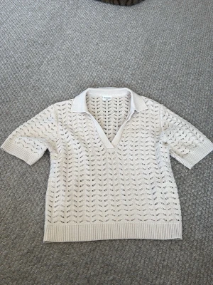 Beige stickad polotopp med krage - Säljer en beige stickad polotopp från Mango med korta ärmar och v-ringning. Toppen har ett snyggt hålmönster och ribbade detaljer vid krage, ärmslut och nederkant. Perfekt för dig som gillar en avslappnad och trendig stil.