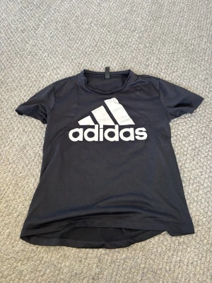 Svart Adidas t-shirt med logga - Snygg svart t-shirt från Adidas med stor vit logga på bröstet. Klassisk rund hals och korta ärmar. Perfekt för dig som gillar sportig stil och vill ha något bekvämt till vardags. Tillverkad i mjuk bomull.