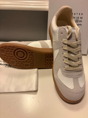Maison Margiela  sneakers - Maison Margiela  sneakers i vitt och beige med ovandel i läder och mocka. Klassisk retrodesign med beige gummisula och matchande snörning. Perfekta för dig som gillar stilrena och tidlösa sneakers med premiumkänsla.