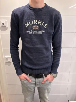 Morris tröja - Snygg blå stickad tröja från Morris med broderad logotyp och brittisk flagga på bröstet. Tröjan har rund halsringning, ribbade muddar och är långärmad. Perfekt för dig som gillar klassisk stil med en twist. M men passar också S.
