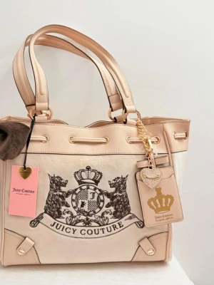 Juicy Couture daydreamer scottie beige  - Stilren beige handväska från Juicy Couture med stort svart logotryck framtill, dekorativ brun rosett på sidan och guldfärgade detaljer. Väskan har dubbla handtag, dragkedja och snygga tags med hjärta och krona. Materialet är en mix av mocka och konstläder.