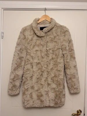 Beige fuskpäls kappa med krage - Snygg beige kappa i fuskpäls med mjuk struktur och klassisk krage. Kappan har knäppning framtill och är helfodrad för extra komfort. Perfekt för dig som vill ha en trendig och mysig look under kalla dagar.
