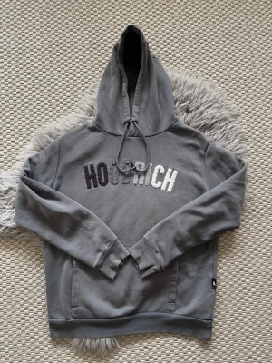 Grå hoodie från Hoodrich med brodyr - Snygg grå hoodie från Hoodrich med broderad logga i olika grå nyanser på bröstet. Hoodien har huva med snörning och långa ärmar. Perfekt för en avslappnad streetwear-look.