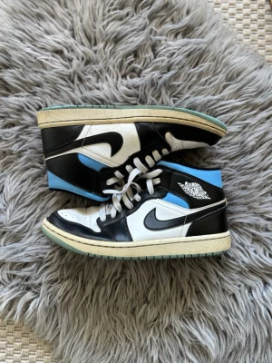 Nike Air Jordan 1 Mid svart/blå/vit - Säljer ett par Nike Air Jordan 1 Mid sneakers i svart, blått och vitt läder. Klassisk siluett med svart swoosh, blå häl och vita paneler. Skön passform och ikonisk Jordan-logga på sidan. Perfekta för dig som gillar streetwear och snygga detaljer.