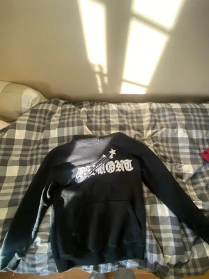 Demont hoodie - Svart hoodie med stort vitt tryck på bröstet där det står 'DEMONT' och ett motiv av stjärnor och linjer. Hoodien har klassisk passform, lång ärm och känguruficka framtill. Perfekt för dig som gillar streetwear och grafiska prints.