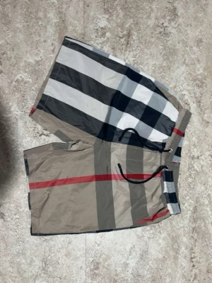 Burberry shorts - Säljer ett par snygga badbyxor med klassiskt rutigt mönster i beige, svart, vitt och rött. De har elastisk midja med svart dragsko och är tillverkade i ett snabbtorkande material. Perfekta för stranden eller poolen. Helt nya aldrig använt 
