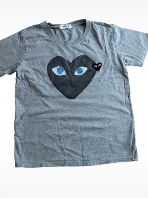 Grå Comme des Garçons Play t-shirt - Grå t-shirt från Comme des Garçons Play med stort svart hjärta och blå ögon på bröstet samt ett litet broderat svart hjärta. Klassisk rund hals och korta ärmar. Perfekt för en cool och avslappnad stil. Tröjan är L men passar M, har du några frågor är det bara att höra av sig!