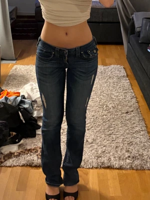 Intressekoll på True religion mörkblå bootcut jeans  - Intressekoll på mörkblå bootcut jeans med låg midja och slitna detaljer framtill. Jeansen har klassisk femficksdesign, silverfärgade knappar och dragkedja. 