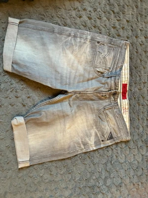 Grå denimshorts från Replay - Snygga grå denimshorts från Replay med klassisk femficksdesign och uppvikta benslut. Shortsen har en avslappnad passform och cool tvättad look, perfekta för en chill stil. Märkesdetalj på fickan och knappgylf.