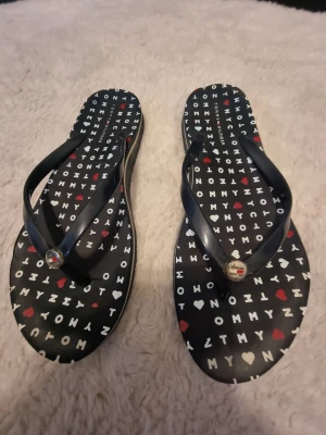 Svarta flip-flops Tommy Hilfiger - Snygga svarta flip-flops från Tommy Hilfiger med bokstäver och röda hjärtan på sulan. Remmarna är i svart plast och har små metalliga detaljer med logga framtill.