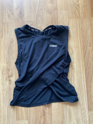 Svart linnetopp från Stronger - Svart ärmlös linnetopp från Stronger med rund halsringning och diskret logga framtill. Toppen har en omlott-detalj framtill och är tillverkad i mjuk bomullsmix, perfekt för träning eller chill. Enkel och stilren design som passar till det mesta. Storlekslappen är bortklippt men skulle säga att den passar S, finns även ett litet hål där.