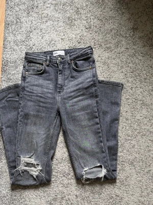Gråa ripped jeans från Zara - Säljer ett par gråa jeans från Zara med hög midja och raka ben. Jeansen har slitningar och hål vid knäna för en trendig, avslappnad look. Klassisk femficksmodell och knappgylf. Perfekta för dig som gillar streetstyle och edgy vibes. Innerbensmått 82,5 cm.