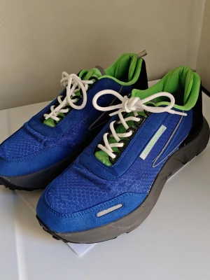 Blå och gröna sneakers från FILA VIBRAM 43 - Snygga sneakers från Fila i blått med gröna detaljer och vit snörning. Skorna har en chunky grå Vibram-sula och hexagonmönstrad mesh på ovandelen. Insidan är limegrön och hälen har en cool grafisk skydd  detalj. Perfekta för dig som gillar streetwear och komfort. Passar löpning , vandring. Kraftiga skor till svår väder.