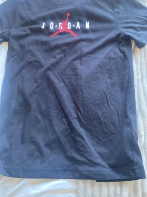 Svart Jordan t-shirt med röd logga - Svart t-shirt från Jordan med klassisk Jumpman-logga i rött och vit text på bröstet. T-shirten har rund halsringning och korta ärmar. Perfekt för dig som gillar sportig och stilren streetwear.