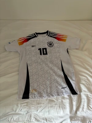 Tysklands fotbollströja Musiala #10 - Säljer en vit Tyskland fotbollströja från Adidas med Musiala och nummer 10 på ryggen. Tröjan har svarta detaljer, tysklands flaggas färger på axlarna och ett tryckt förbundsmärke på bröstet. Tillverkad i lätt och ventilerande material, perfekt för match eller träning.