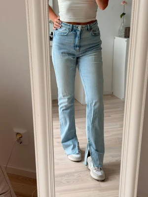 Ljusblå raka jeans med slits - Säljer ett par ljusblå jeans med raka ben och hög midja. Jeansen har en snygg slits längst ner vid bensluten och rå kant. Klassisk femficksmodell och stängs med dragkedja och knapp. Perfekta till sneakers och crop tops. Har tyvärr fläckar på benet, se bild.