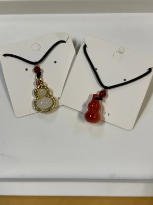 Två halsband med jade kinesisk kalebass - Säljer två halsband med svarta band och jade kalebassformade hängen. Ett halsband har ett vitt jade hänge med guldfärgad ram och gnistrande stenar, det andra har ett rött hänge i jade. Båda har små röda pärlor ovanför hänget. Perfekta för dig som gillar unika detaljer.