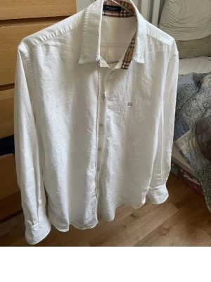 Vit skjorta från Burberry med rutiga detaljer - Klassisk vit skjorta från Burberry med ikoniska beige-rutiga detaljer på insidan av kragen och manschetterna. Skjortan har knappar framtill, lång ärm och en liten broderad logga på bröstet. Perfekt för en stilren och trendig look.