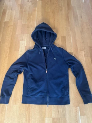 Mörkblå hoodie från Morris - Snygg mörkblå hoodie från Morris med dragkedja, huva med dragsko och två fickor framtill. Broderad logga på bröstet och ribbade muddar vid ärmslut och nederkant. Perfekt för en chill och stilren look. Bra skick inga stora defekter förutom ett litet hål vid slutet av armen på hoodien, ny pris ligger runt 1200kr
