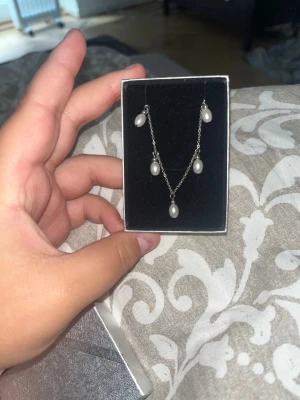 Silverhalsband med vita pärlor - Snyggt halsband i silverfärgad metall med fem droppformade vita pärlor. Kedjan är tunn och elegant, och pärlorna hänger i olika nivåer för en modern look. Perfekt accessoar för att lyfta din outfit. Nypriset är 420 kr