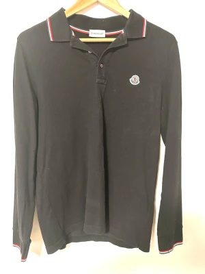 Moncler Långärmad Piké | M - Snygg svart långärmad pikétröja från Moncler med klassisk krage och knäppning. Använd fåtal gånger därav i väldigt fint skick