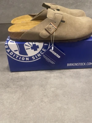 Birkenstock Boston taupe tofflor - Säljer ett par Birkenstock Boston tofflor i färgen taupe. De är tillverkade i mjukt mockaläder med justerbart spänne i metall och har en klassisk korksula för extra komfort. Perfekta för dig som gillar stilrena och bekväma skor.