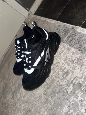 Svarta sneakers från DlOR - Snygga sneakers från DlOR i svart med vita detaljer. Skorna har en kraftig sula, meshpartier för extra ventilation och reflekterande inslag längs sidorna. Klassisk snörning och sportig look som passar till streetwear och casual outfits.